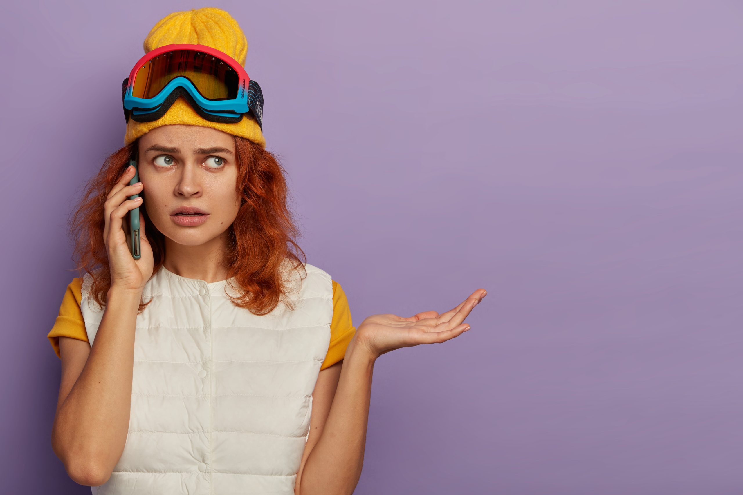 puzzled dissatisfied woman spends free time on ski resort, phones in roaming, holds mobile phone near ear, raises palm, cannot solve problem, poses against purple wall, blank space. winter season